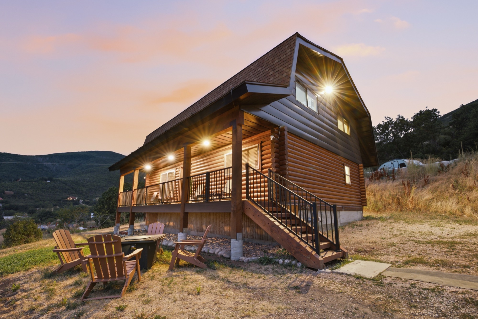 A log cabin at dusk with porch lights on and Adirondack chairs by a fire pit, managed by Vanik