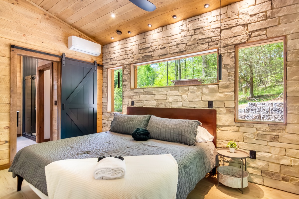 A stone-walled cabin bedroom staged by Vanik with picture windows into the forest